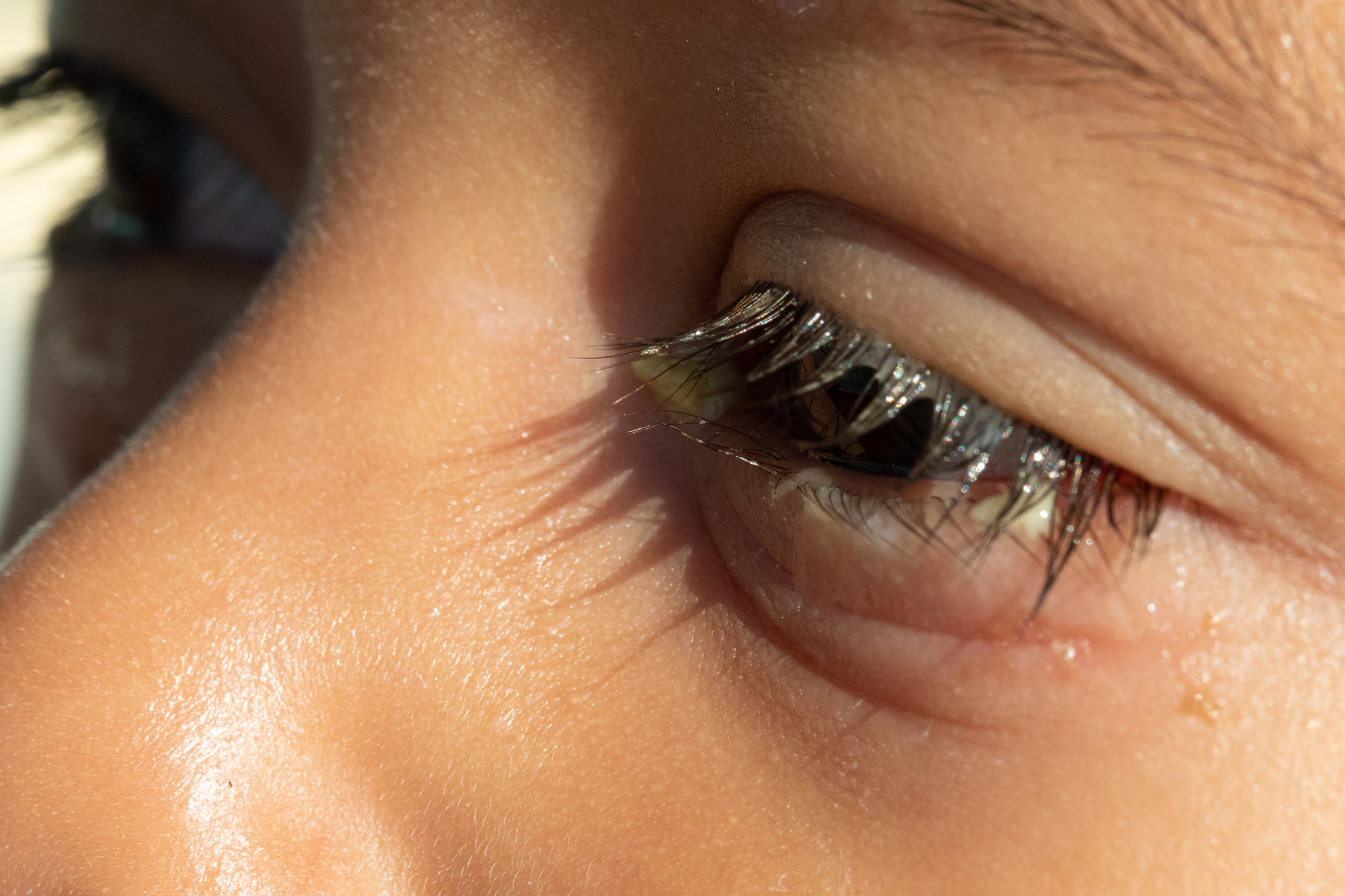 Close-up of a person's eye with long eyelashes, showing details of the eyelid and surrounding skin. The side view captures the natural texture and lighting on the skin and lashes.
