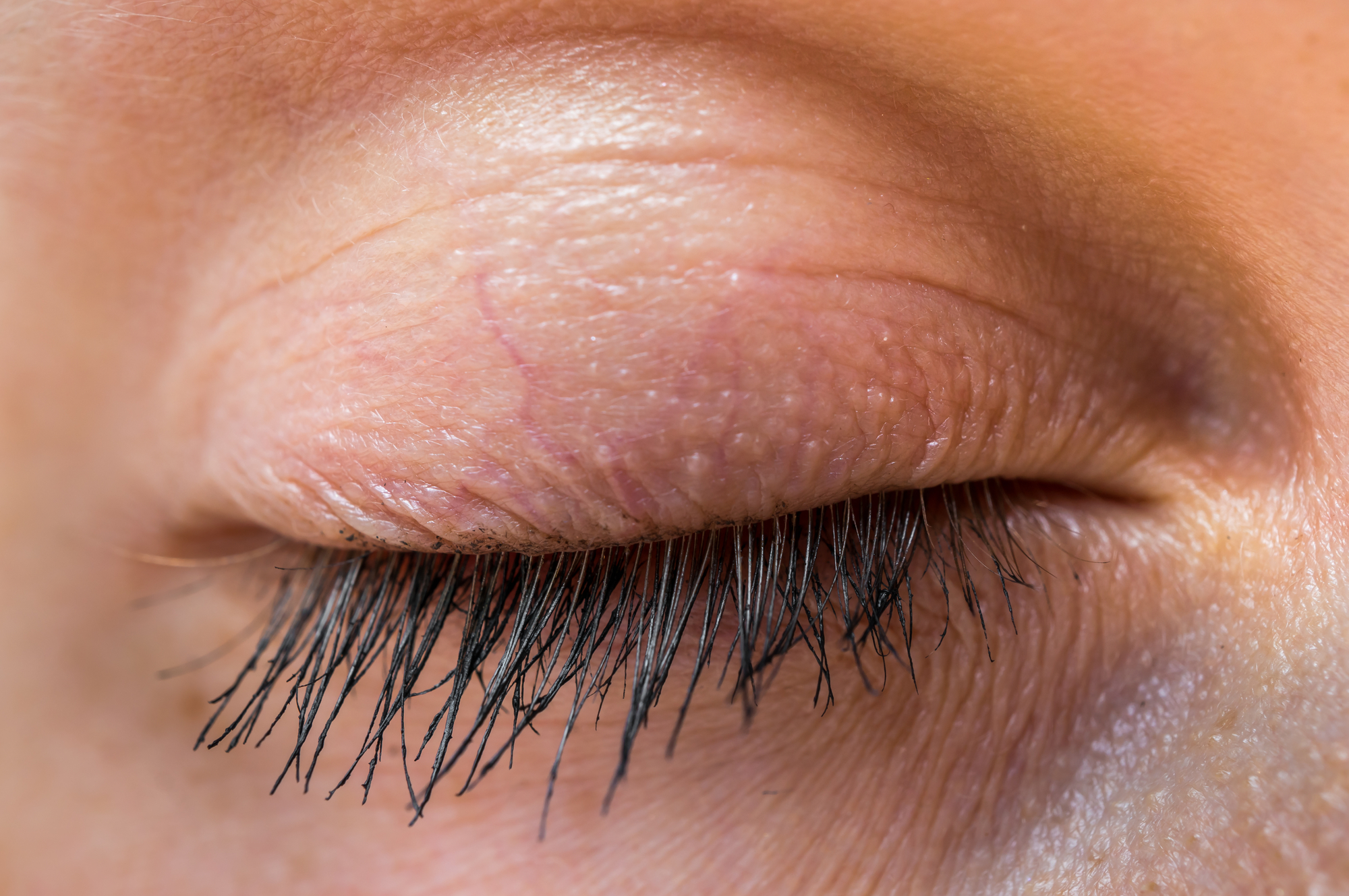 Close-up of a closed human eye with long eyelashes. The skin around the eye has a natural texture, and faint veins are visible on the eyelid. The lighting highlights the details of the eyelashes and skin.